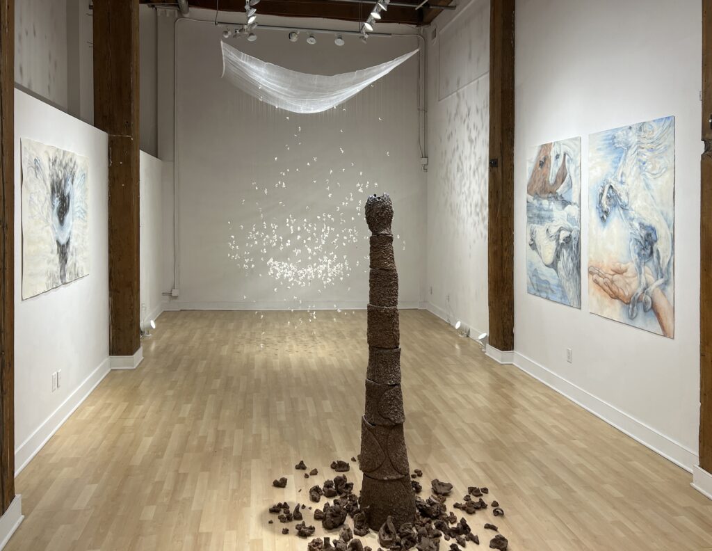 Gallery installation with brown tower with crumbled pieces at the base, and white fabric and many small pieces of white scraps hanging below