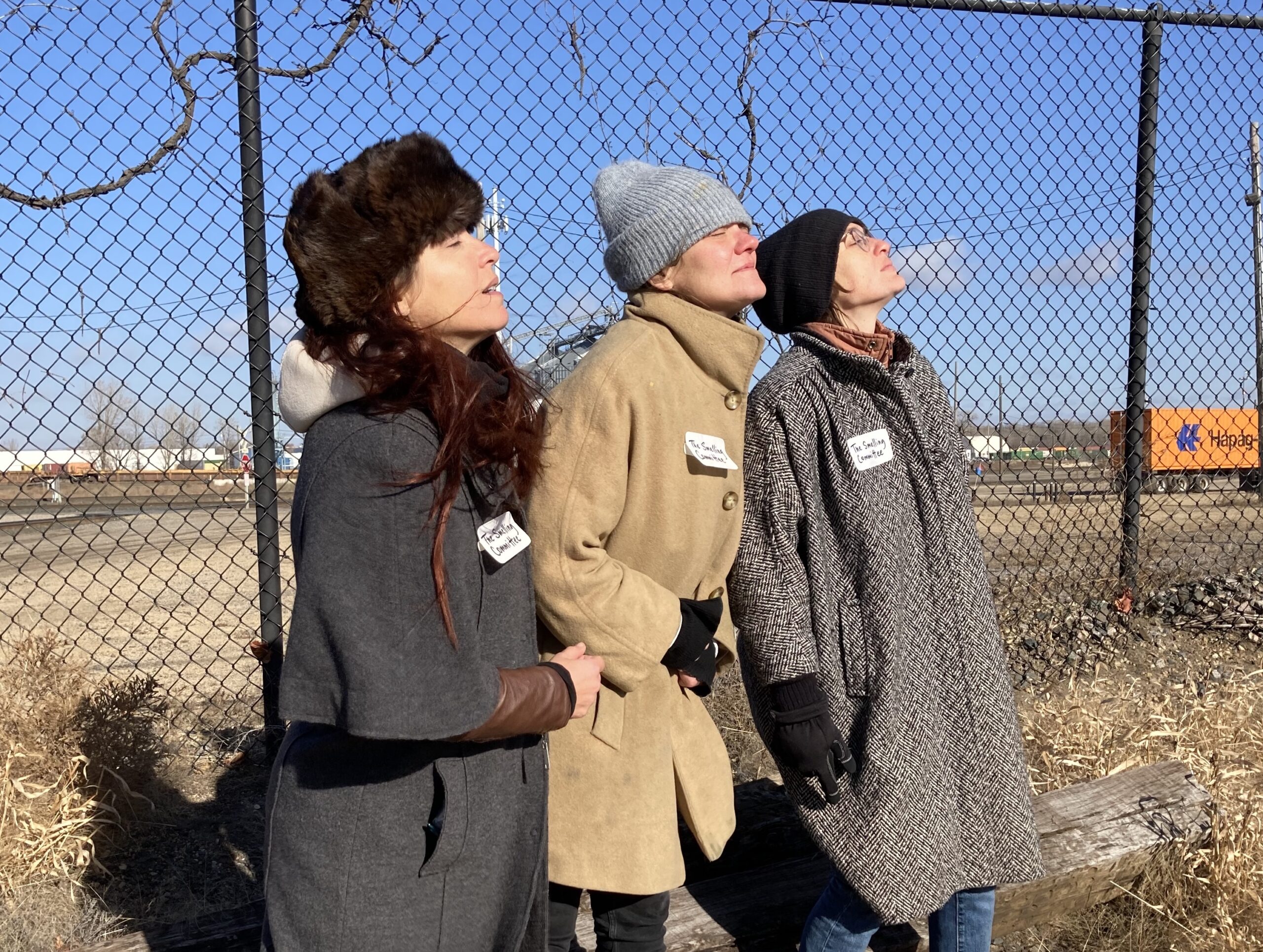 Three people stand in front of fence, eyes closed, noses upturned to smell